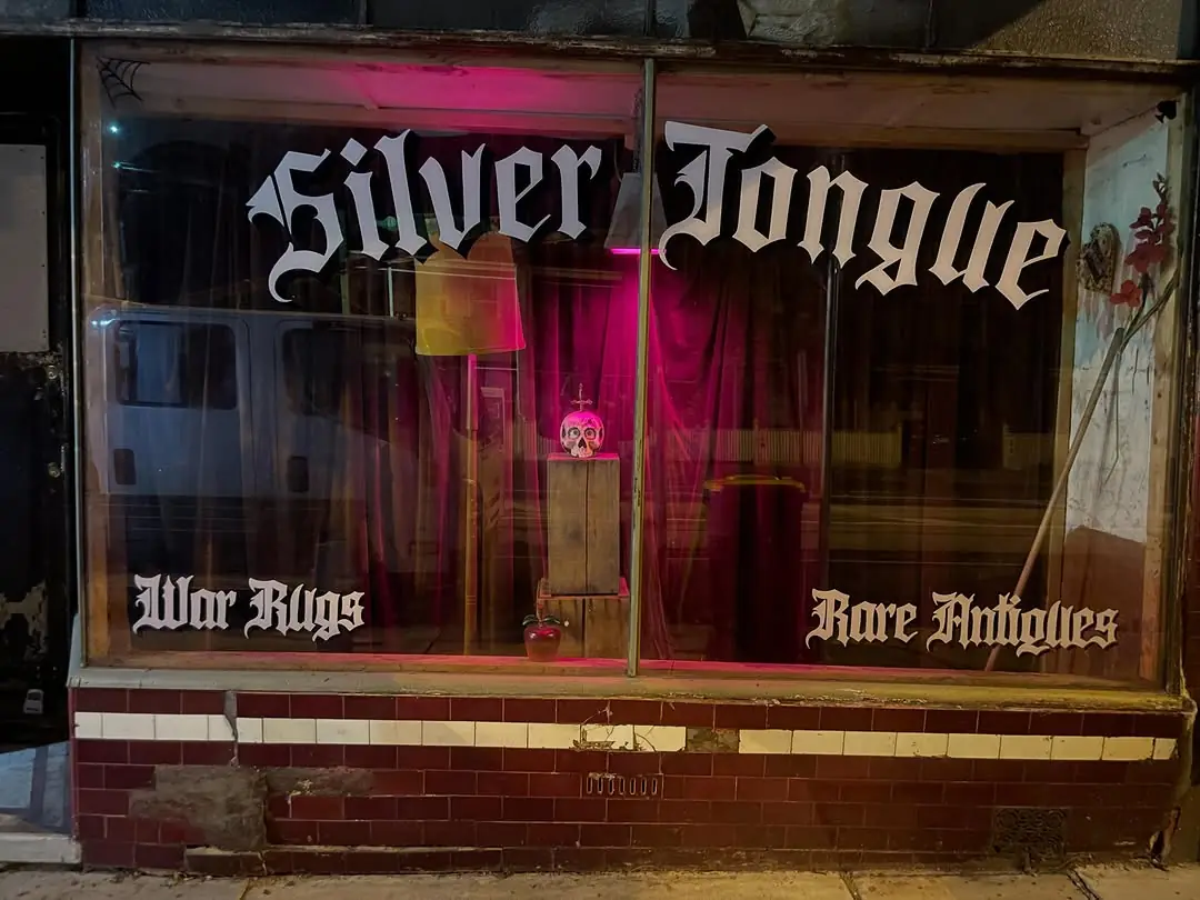 The Silver Tongue shop window at night, lettered War Rugs and Rare Antiques